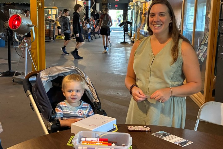 Woman smiling beside child in stroller with art supplies on table.