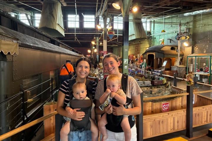 Two adults with babies in carriers standing inside a train museum.