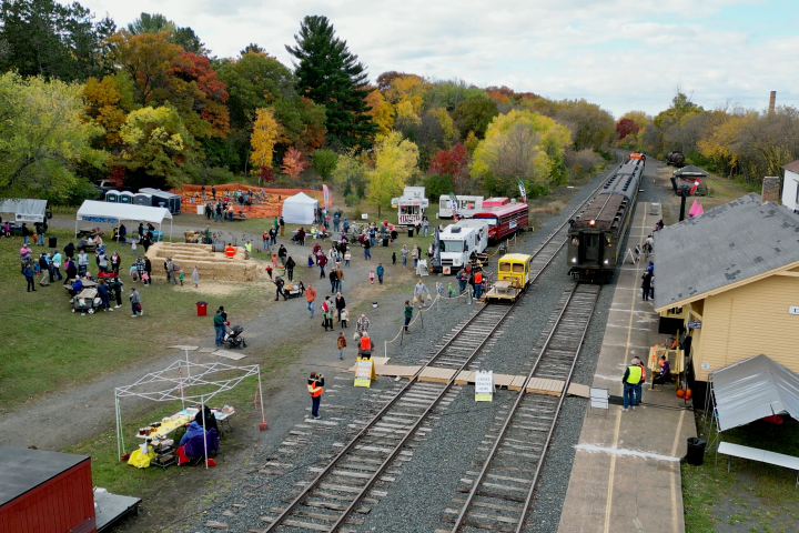 Autumn festival with booths, crowds, and train at rural station among colorful trees.