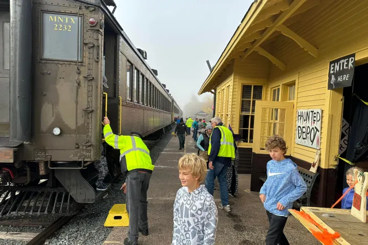 People boarding a vintage train at a station with a yellow building.