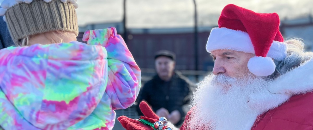 Santa Claus with bells greets child in colorful jacket and cupcake hat outdoors.
