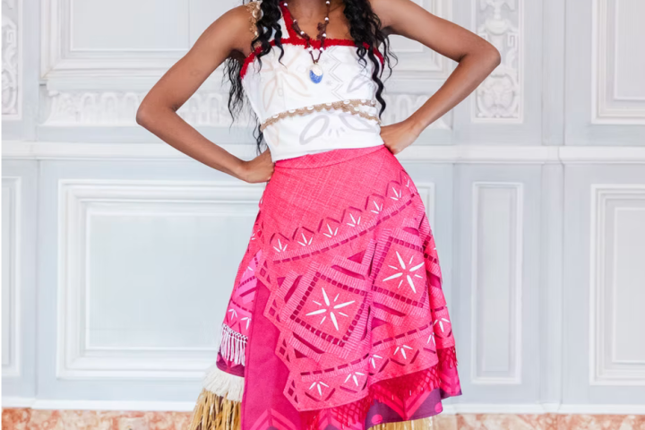 Person in Polynesian outfit with red skirt, grass details, and floral headpiece, standing indoors.