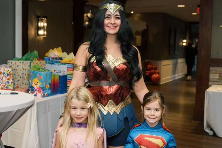 Woman in superhero costume with two kids holding superhero posters at a party.